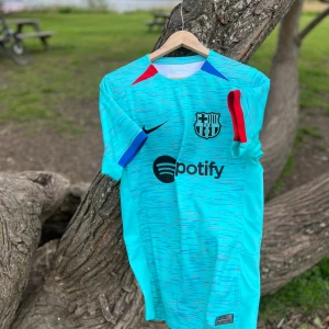 FC Barcelona Tredjetröja 2023/2024 - Säljer en turkos FC Barcelona fotbollströja från Nike med svarta, blå och röda detaljer, samt klubbmärke och sponsortryck på bröstet. Tröjan är kortärmad och tillverkad i Dri-FIT-material som andas. .