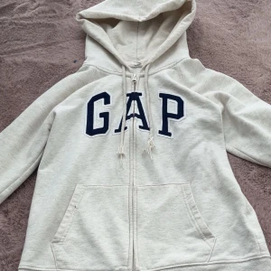 Beige hoodie med dragkedja från GAP - Säljer en beige hoodie från GAP med dragkedja framtill och stora fickor. Tröjan har huva med snören och GAP-logga i mörkblått på bröstet. Perfekt för en avslappnad stil.