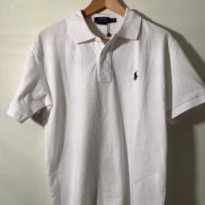 Ralph Lauren Piké - Klassisk vit piké t-shirt från Polo Ralph Lauren med korta ärmar och broderad logga på bröstet. Tidlös design med krage och knappar framtill. Perfekt för en clean och stilren look.