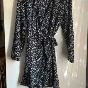 Marinblå blommig omlottklänning med volang - Söt marinblå omlottklänning med vitt blommönster och volangdetaljer längs ringningen och fållen. Klänningen har lång ärm och knyts i midjan. 