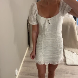 Vit kortklänning med puffärm från NA-KD - Superfin vit kortklänning från NA-KD med broderade blommor och puffärmar. Klänningen har knytning framtill, v-ringning och är figurnära i modellen. Perfekt för sommarens alla tillfällen!