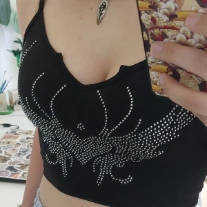 Svart croppad linne-topp med strasshjärta - Säljer en svart croppad linne-topp med smala axelband och ett hjärtmönster i silvriga strasstenar framtill. Toppen har en liten v-ringning och är en bra statementpiece.