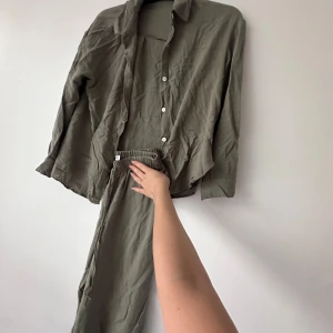 Olivgrön skjorta med knappar och byxor - Säljer en olivgrön skjorta med klassisk krage och vita knappar framtill. Skjortan har lång ärm och är tillverkad i ett mjukt, tunt tyg som känns skönt mot huden. Perfekt att styla med matchande byxorna