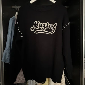 Svart stickad tröja med texten 'Magical' - Svart stickad tröja från MacyMccoy med vit broderad text 'Magical' på bröstet. Tröjan har långa ärmar, ribbade muddar och slitna detaljer vid fållen och ärmsluten samt vita stygn längs axlarna.
