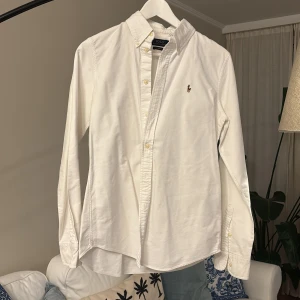Vit skjorta från Polo Ralph Lauren - Klassisk vit skjorta från Polo Ralph Lauren med broderad logga på bröstet. Skjortan har lång ärm, knappar framtill och button-down krage. Perfekt för en clean och stilren look.