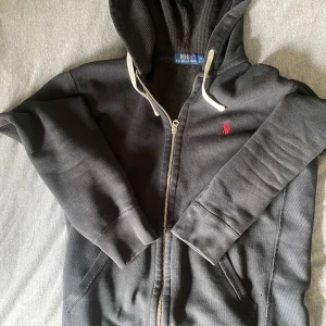Svart hoodie från Polo Ralph Lauren - Säljer en svart hoodie från Polo Ralph Lauren med dragkedja och huva. Tröjan har en röd broderad logga på bröstet och vita snören i huvan. Perfekt för en avslappnad och stilren look.
