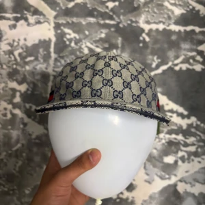 Gucci keps med monogram och röd-blå rand - Snygg keps från Gucci med klassiskt GG-monogram i blått på beige botten. Kepsen har en röd och blå rand på sidan och justerbar rem baktill. Materialet är bomull och kepsen har böjd skärm. Storlekn är L passar mitt huve perfekt har måtten 57 cm. PRISET ÄR INGE HUGGET I STEN!