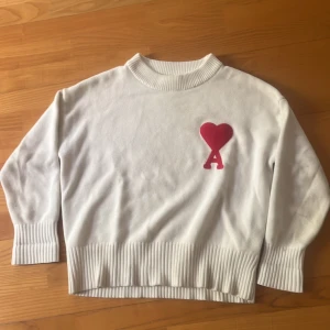 Vit stickad tröja från Ami Paris med rött hjärta - Vit stickad tröja från Ami Paris med ribbade muddar och nederkant. På bröstet finns ett rött hjärta med bokstaven A. Tröjan har lång ärm och en klassisk rund halsringning. Perfekt för en stilren och trendig look.