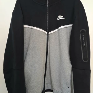 Nike zip-hoodie i svart och grå - Snygg zip-hoodie från Nike i svart och grå med vit logga på bröstet. Jackan har dragkedja framtill, huva och en ficka med dragkedja på ärmen. Perfekt för träning eller chill.