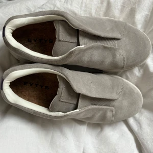Gråa mockasneakers från Eytys - Säljer ett par stilrena sneakers från Eytys i beige grå med vit sula och diskret lila detalj bak. Skorna har en minimalistisk design utan synlig snörning och är fodrade med vitt tyg invändigt. Perfekta för dig som gillar clean och modern stil.                                                             Dom är i väldigt bra skick då de används ett fåtal gånger sen de köptes i april. Säljer dom för att dom inte kommer till användning.