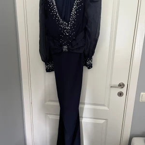 Marinblå långklänning med pärldetaljer från SHEN Belle - Elegant marinblå långklänning med långa puffärmar och v-ringning dekorerad med pärlor. Klänningen har även pärldetaljer vid ärmsluten och en figurnära passform som ger en lyxig känsla.