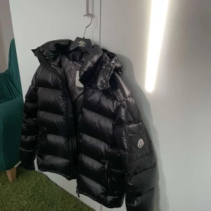 Svart dunjacka från Moncler - Säljer en svart, glansig dunjacka från Moncler med huva och dragkedja. Jackan har en klassisk pufferstil och Moncler-logga på ärmen. Perfekt för kalla dagar och riktigt snygg till streetwear.