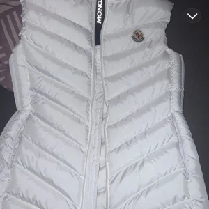 Vit dunväst från Moncler - Snygg vit dunväst från Moncler med dragkedja framtill och klassisk Moncler-logga på bröstet. Västen har en quiltad design och är perfekt för lager-på-lager-stil under kyliga dagar.