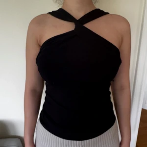 Svart no shoulder topp med twistad hals - Säljer en svart ribbad topp med unik twistad hals och no shoulder-design. Toppen är tight och har en stilren look som passar perfekt till många olika outfits.