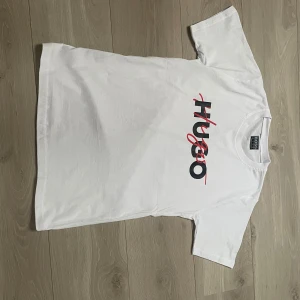 Vit t-shirt från Hugo med logotyp - Vit t-shirt från Hugo med tryckt logotyp i svart och rött på bröstet. Klassisk rund halsringning och korta ärmar. Perfekt för en stilren och enkel look.