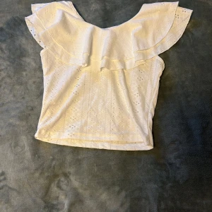 Vit blus med volang - Säljer en söt vit blus med bred volang runt axlarna och v-ringad hals. Blusen har broderade detaljer och är ärmlös, perfekt för varma dagar. Passar till både jeans och kjol för en fräsch look. Den är aldrig använd 