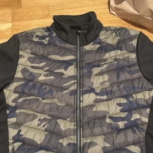 Camouflageväst från Peak Performance - Snygg väst från Peak Performance med camouflage-mönster i gröna och blå toner. Västen har dragkedja framtill och hög krage. Materialet är en blandning av syntet och polyester, vilket gör den lätt och bekväm.