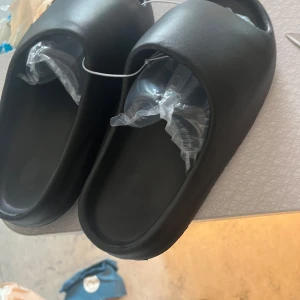 Svarta Yeezy slide  - Säljer ett par svarta chunky sandaler med öppen tå och platt sula. De har en minimalistisk design och är tillverkade i syntetmaterial. Perfekta för sommaren och riktigt sköna att glida runt i.