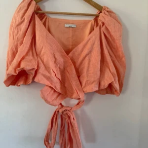 Persikofärgad omlottopp från Gina Tricot - Säljer en persikofärgad omlottopp från Gina Tricot i storlek 38. Toppen har puffiga ärmar, knytning framtill och är tillverkad i ett lätt linnematerial. Perfekt för vår och sommar!