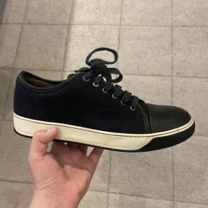 Lanvin cap toe  - Säljer ett par svarta sneakers från Lanvin med vita sulor och svarta snören. Skorna har rund tå, platt sula och detaljer i både tyg och skinn. Klassisk och stilren design med metallöglor för snörningen.