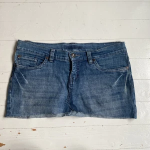 Lågmidjad jeanskjol med utsmyckade bakfickor - Säljer en blå lågmidjad jeanskjol med rå kant nertill och snygga utsmyckade detaljer på bakfickorna. Kjolen har klassisk femficksdesign, bälteshällor och knappgylf. Perfekt för dig som gillar denim med en twist. Midjemått: ca 43cm(rakt över)  grenhöjd: 24cm.   Bakre längd: 30cm