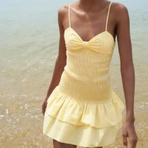 Gul rutig volangklänning med rosett från Zara  - Från Zara. Supersöt gul och vit rutig klänning med smala axelband, volangkant och stor rosett framtill. Klänningen har öppen rygg och omlottkänsla baktill. Perfekt för varma dagar och somriga tillfällen. Använt 1 gång tidigare