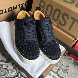  Christian Louboutin - Säljer ett par mörkblåa sneakers från Christian Louboutin med ikoniska röda sulor och svarta nitar på tån. Skorna har snörning och en platt vit sula. Materialet är en mix av mocka och textil, vilket ger en lyxig känsla.