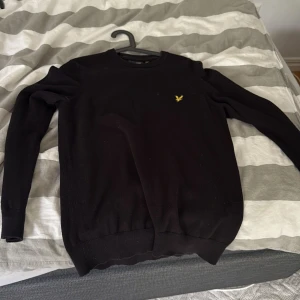 Svart stickad tröja från Lyle & Scott - Säljer en svart stickad tröja från Lyle & Scott med deras klassiska gula fågel-logga på bröstet. Tröjan har rund halsringning och långa ärmar, perfekt för lager-på-lager under kallare dagar.