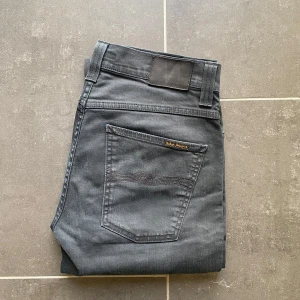 Nudie Jeans Slim Jim - Säljer ett par mörkgrå jeansbyxor från Nudie med klassisk femficksdesign och raka ben. Byxorna har Nudie-logga på bakfickan och svart patch bak i midjan. Perfekta för dig som gillar en enkel och stilren look.