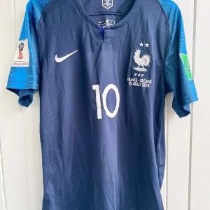 Kylian Mbappe - Frankrike VM-final 2018 - Säljer en klassisk fotbollströja, Kylian Mbappes matchtröja från VM-finalen 2018 när han var med och avgjorde så att hans Frankrike vann över Kroatien och blev världsmästare. Tröjan är en remake i nyskick. världsmästare. Tröjan är en remake i nyskick och har finalen och datumet inskrivet på bröstet (samma text som Mbappe hade på sin tröja under finalen). Aldrig använd.  Notera: Tröjan har en logo i nacken upp och ner (se sista bild). Synd knappt dock.  Large, normal i storleken.  Skickas inom 24