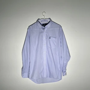 Ljusblå randig skjorta från Ralph Lauren - Klassisk ljusblå skjorta från Ralph Lauren med tunna vita ränder och broderad logga på bröstfickan. Skjortan har knappar framtill, lång ärm och krage. Perfekt för en stilren look.