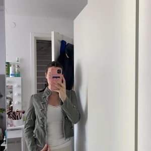 Olivgrön militärinspirerad jacka med knappar - Snygg olivgrön jeans jacka i militärstil med dekorativa knappar framtill och på axlarna. Jackan har ståkrage och markerad midja för en cool och trendig look. Perfekt att styla till jeans eller kjol.