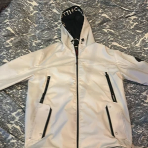 Vit jacka från Zavetti Canada - Säljer en snygg vit jacka från Zavetti Canada med svarta detaljer och dragkedjor. Jackan har huva med logotyp, två framfickor med dragkedja och en patch på ärmen. Perfekt för dig som vill sticka ut med stilren design.