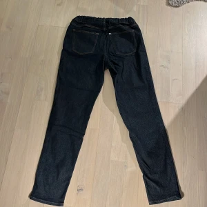 Mörkblå jeansbyxor med resårmidja - Säljer ett par mörkblå jeansbyxor med resår i midjan och klassiska bakfickor. Byxorna har raka ben och stängs med knapp och dragkedja framtill. Perfekta för en avslappnad och stilren look.