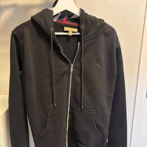 Svart hoodie  från BB med dragkedja - Svart hoodie med klassiskt rutigt foder i huvan och broderad logga på bröstet. Tröjan har dragkedja framtill, snörning i huvan och två stora fickor. Perfekt för dig som gillar stilrena plagg.