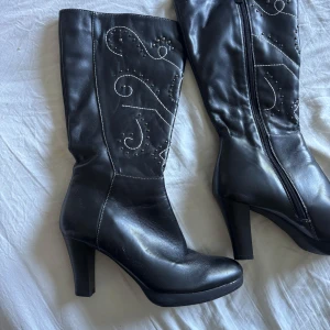 Svarta stövlar med klack och broderi - Snygga svarta stövlar i skinn med hög klack och rund tå. Dekorerade med vita broderade mönster och små nitar längs skaftet. Dragkedja på insidan för enkel påtagning. sparsamt använda men har lite jack på men syns knappt🩷