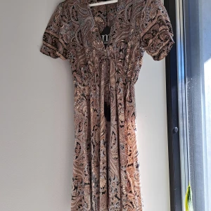 Mönstrad klänning Manoukian S - Snygg klänning från Manoukian med korta ärmar och v-ringning. Klänningen har ett paisleymönster i beige, brun och svart, samt knytning under bröstet. Tillverkad i 100% viskos för en luftig känsla. Otroligt snyggt material som liknar silke (inte genomskinlig). Ny med lappen kvar. Fransk designer.