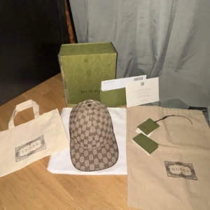 Äkta gucci keps  - Exklusiv Gucci keps i beige med klassiskt GG-monogram över hela kepsen. Baksidan har den ikoniska gröna och röda randen. Justerbar rem baktill i brunt läder. Kepsen är tillverkad i canvas och levereras med originalbox, dustbag och tags.