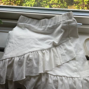 Vit volangkjol från NLY Trend - Supersöt vit kortkjol från NLY Trend i storlek S. Kjolen har volanger och en lätt skrynklig struktur som ger en chill och trendig vibe. Perfekt för varma dagar och snygg att styla med crop tops eller oversized t-shirts.