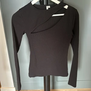 Svart ribbad topp med cut out NLY Trend - Svart långärmad topp från NLY Trend i ribbad bomullsmix. Snygg cut out-detalj vid axeln och asymmetrisk halsringning som ger en trendig look. Perfekt för dig som vill sticka ut lite extra.