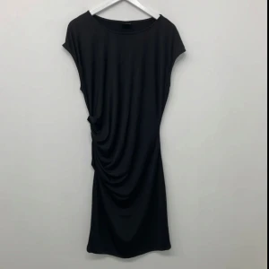 Svart draperad kortklänning - En svart kortklänning med snygg drapering på ena sidan. Klänningen har rund halsringning och är ärmlös, vilket ger en clean och modern look. Perfekt för dig som gillar minimalistisk stil med en twist. Materialet känns mjukt och följsamt mot kroppen.