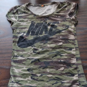 Nike camo t-shirt med stor logga - Camo t-shirt från Nike i gröna och bruna toner med stor svart Nike-logga på bröstet. Klassisk rund hals och korta ärmar. Perfekt för dig som gillar streetstyle och sportiga vibes.