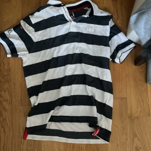 Randig pikétröja från Helly Hansen - Snygg pikétröja från Helly Hansen i svart och vitt med breda ränder. Klassisk krage, kort ärm och knäppning vid halsen. Perfekt för en sportig och avslappnad stil. Stora loggor på ärm och bröst ger en cool touch.