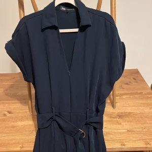 Jumpsuit med bälte - Snygg mörkblå jumpsuit från Zara med krage, korta ärmar och markerad midja med brett bälte och metallspänne. Vida ben och dolda knappar framtill. Perfekt för dig som gillar stilrena och bekväma plagg med en modern touch.