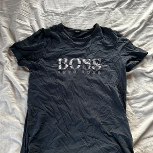 Svart t-shirt från Hugo Boss - Mörkblå t-shirt från Hugo Boss med stort BOSS-tryck på bröstet där bokstäverna har ett färgglatt mönster inuti. Klassisk rund halsringning och korta ärmar. Skön och mjuk bomullskvalitet, storlek m