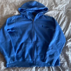 Blå hoodie från Polo Ralph Lauren - Snygg blå hoodie från Polo Ralph Lauren med dragkedja framtill och klassisk broderad logga på bröstet. Tröjan har huva, kängurufickor och är tillverkad i mjuk bomull. Perfekt för en chill och avslappnad stil. Det finns ett hål på luvan men som ej är märkvärdigt, går även att fixa. 