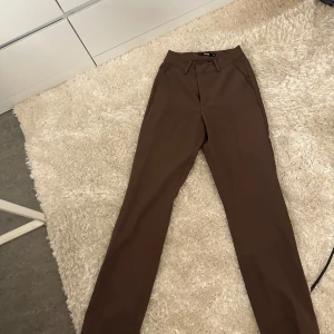 Bruna kostymbyxor med raka ben - Snygga bruna kostymbyxor från NA-KD med raka ben och klassisk passform. Byxorna har hällor för bälte och sidfickor, perfekta för en clean och stilren look. Materialet känns mjukt och har en lätt stretch för extra komfort.