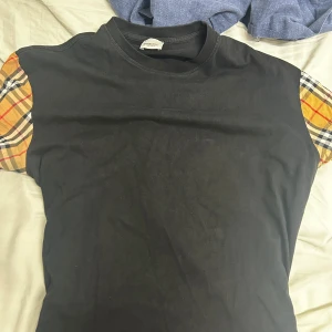 Svart t-shirt med rutiga ärmar Burberry - Svart t-shirt från Burberry med klassiska rutiga korta ärmar i beige, rött och svart. T-shirten har en enkel rund halsringning och är tillverkad i mjuk bomull. Perfekt för dig som vill sticka ut med en unik twist på en basic tee.