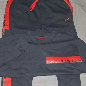 Svart tröja från Hugo med röd logga - Svart långärmad tröja från Hugo med röd logga och tryck längs ena ärmen. Tröjan har rund halsringning och ribbade muddar. Snygg kontrast med rött tryck och detaljer, perfekt för en streetwear-look.