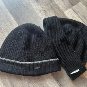 2 svarta mössor BOSS & Tiger of Sweden - Två stilrena svarta ribbstickade mössor, en från BOSS med grå rand och metallogo framtill, och en från Tiger of Sweden med diskret etikett. Båda är i mjuk ullblandning och passar perfekt för kalla dagar.